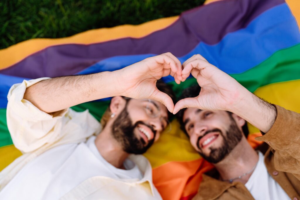 Happy young male couple lying on rainbow flag and showing heart gesture with hands
