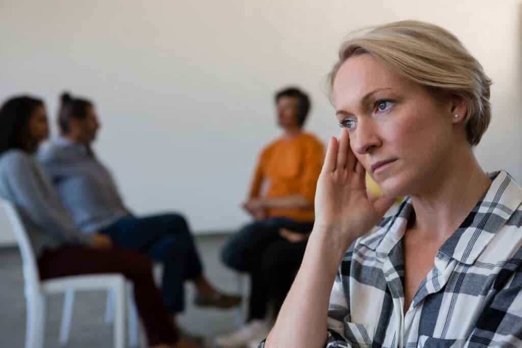 A woman with blonde hair and a checkered shirt rests her head on her hand and looks off to the side